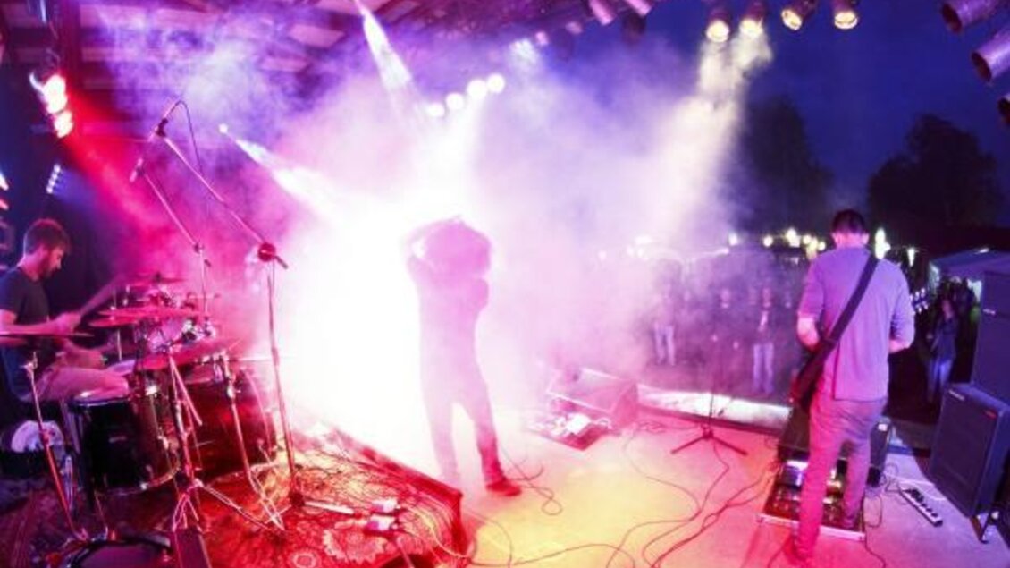 Eine Band spielt auf einer Bühne mit bunten Lichtern und Nebel. Die Menge schaut begeistert zu.