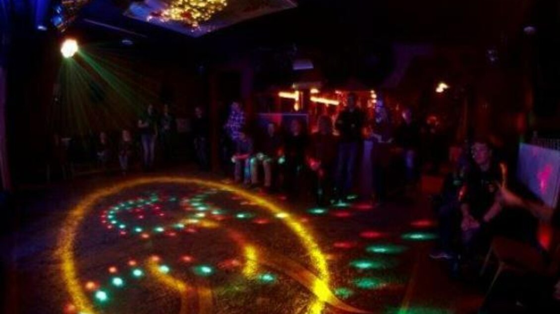 Eine lebhafte Tanzfläche mit bunten Lichtern und Schatten. Im Hintergrund sind Menschen zu sehen, die Spaß haben.