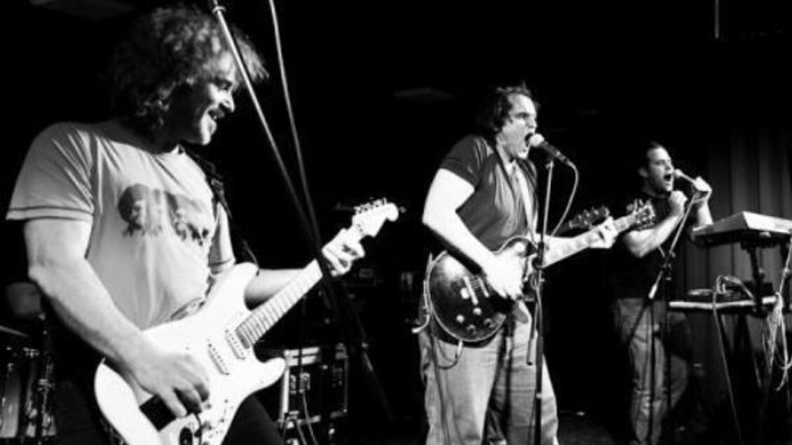 Eine Band spielt live auf der Bühne. Die Musiker sind leidenschaftlich aktiv und nutzen verschiedene Instrumente.