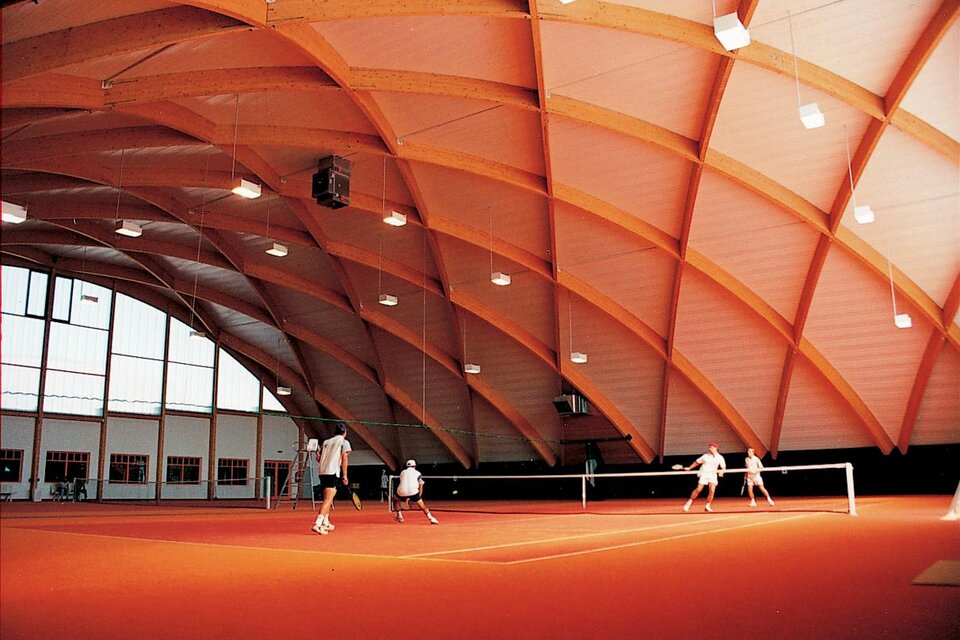 Eine moderne Tennishalle mit einem hohen, gewölbten Dach. Auf dem leuchtend orangefarbenen Platz spielen mehrere Personen Tennis. | © Lederer