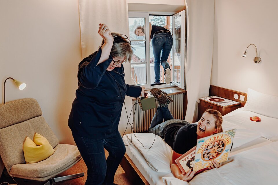 Eine humorvolle Szene in einem Hotelzimmer, in der eine Frau fröhlich im Bett liegt und ein Buch anschaut, während eine andere Frau durch das Fenster klettert. Die Atmosphäre ist entspannt und lustig. | © Der Eberhard