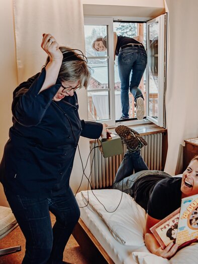 A humorous scene in a hotel room, where a woman is cheerfully lying in bed looking at a book, while another woman is climbing through the window. The atmosphere is relaxed and funny. | © Der Eberhard