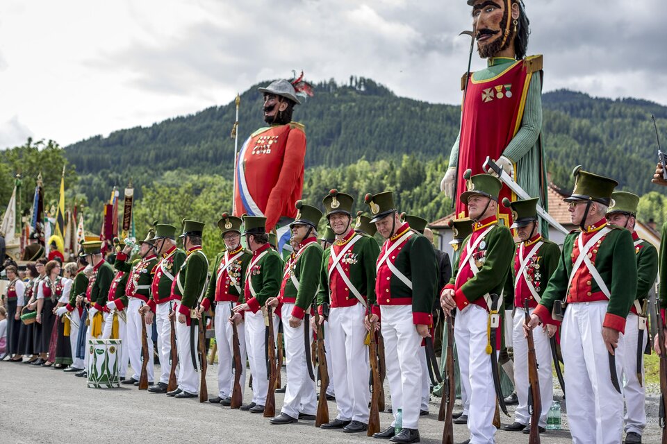 Ein festlicher Umzug mit Menschen in historischen Uniformen und großen Figuren. Im Hintergrund sind grüne Hügel und Wolken sichtbar. | © Steiermark Tourismus, ikarus.cc