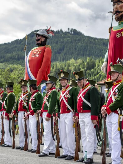A festive parade with people in historical uniforms and large figures. In the background, green hills and clouds are visible. | © Steiermark Tourismus, ikarus.cc