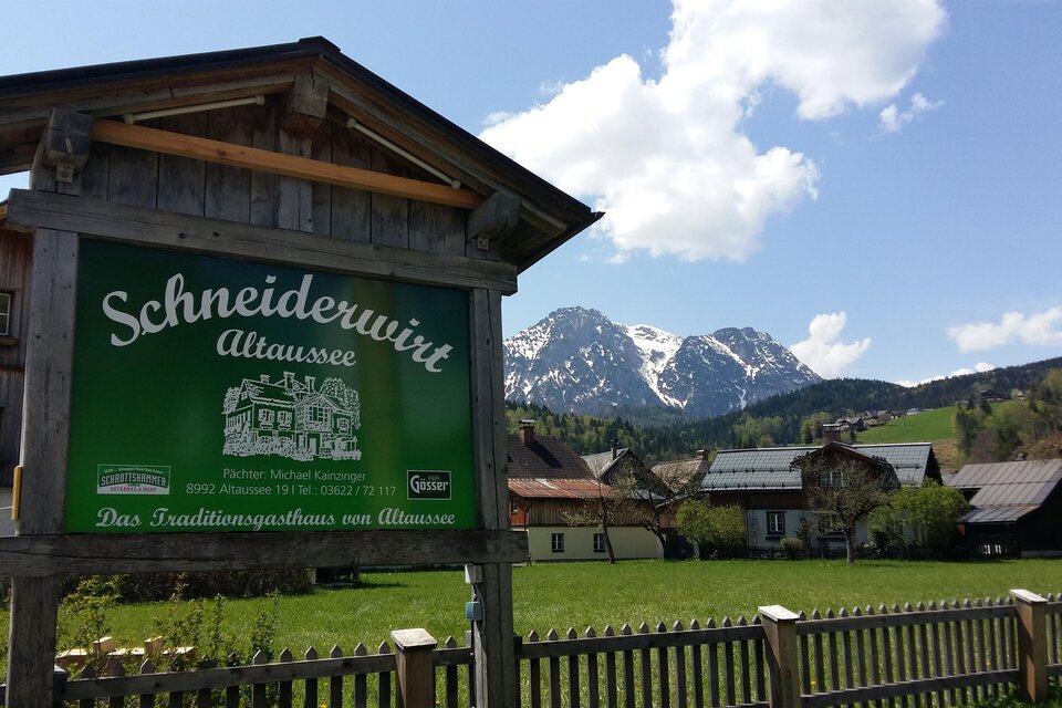 Ein traditionelles Gasthaus mit einem grünen Schild, umgeben von einer malerischen Landschaft. Im Hintergrund sind schneebedeckte Berge und idyllische Bauernhäuser zu sehen. | © Petra Kirchschlager