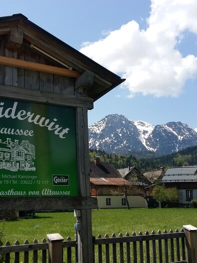 Ein traditionelles Gasthaus mit einem grünen Schild, umgeben von einer malerischen Landschaft. Im Hintergrund sind schneebedeckte Berge und idyllische Bauernhäuser zu sehen. | © Petra Kirchschlager