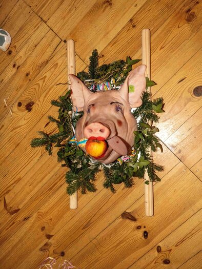 An artistically decorated pig's head with an apple in its mouth, surrounded by green branches. The background is a wooden floor. | © Musikverein Stubenberg