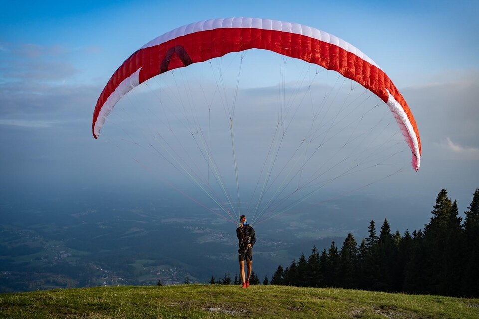 Ein Paraglider steht auf einer grünen Wiese mit einem roten Schirm über sich. Im Hintergrund sind Bäume und ein bewölkter Himmel zu sehen. | © @Steinbauer