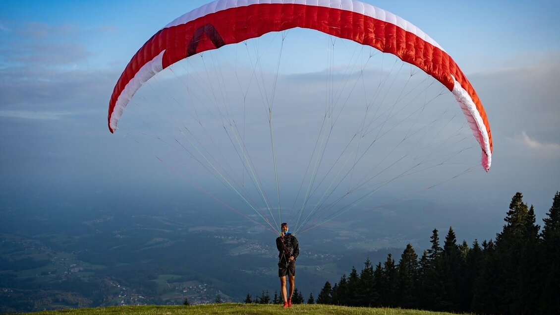 Ein Paraglider steht auf einer grünen Wiese mit einem roten Schirm über sich. Im Hintergrund sind Bäume und ein bewölkter Himmel zu sehen. | © @Steinbauer