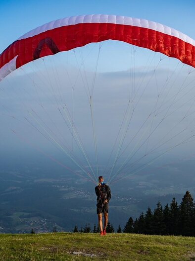 Ein Paraglider steht auf einer grünen Wiese mit einem roten Schirm über sich. Im Hintergrund sind Bäume und ein bewölkter Himmel zu sehen. | © @Steinbauer