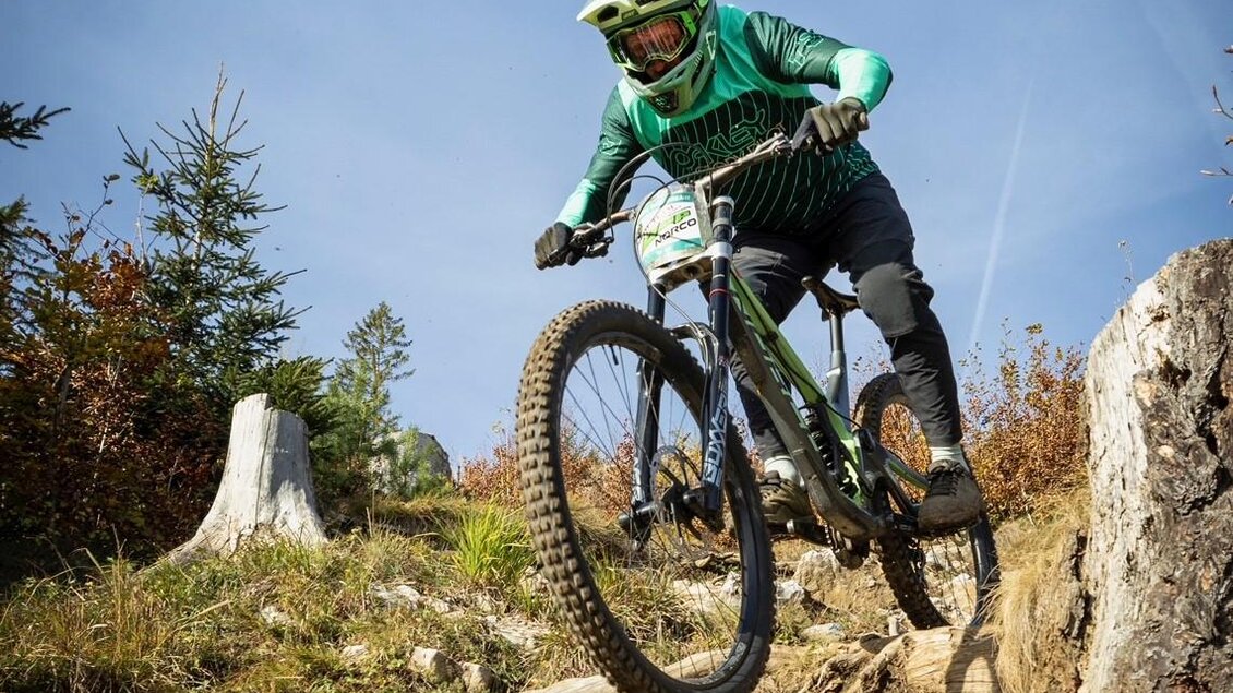 Ein Mountainbiker springt über einen Felsen in der Natur. Die Umgebung ist grün und bewaldet, mit klarem Himmel. | © @Steinbauer