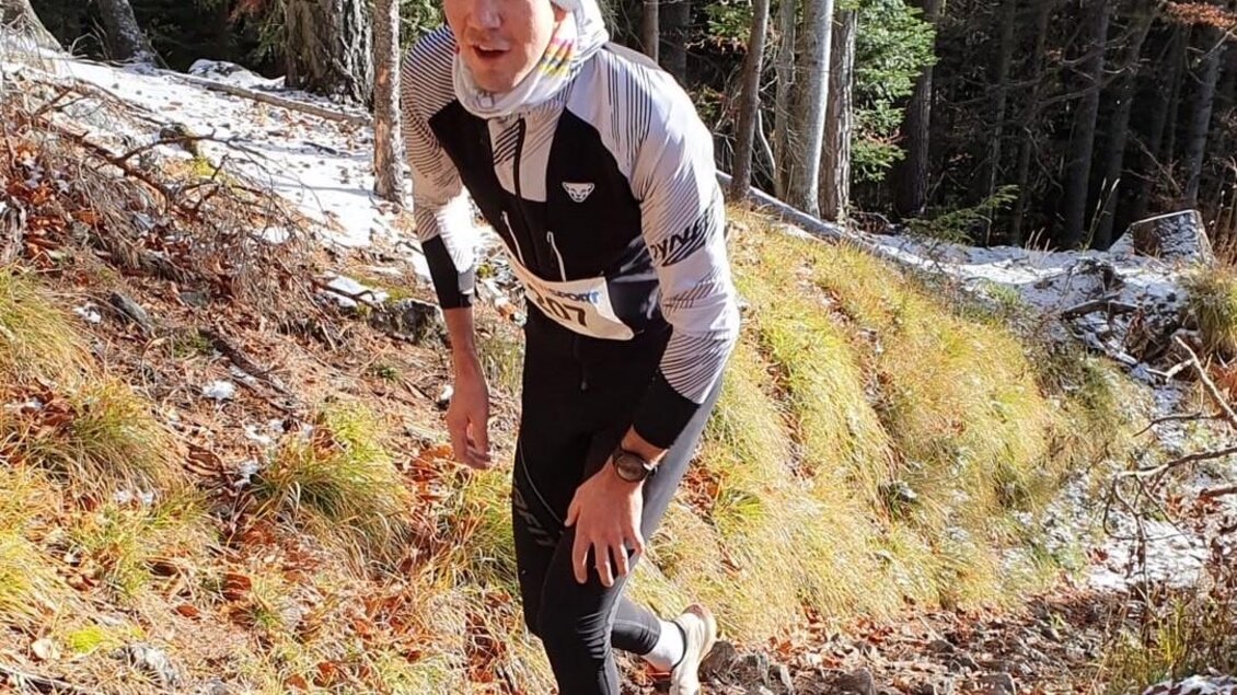 Ein Läufer in sportlicher Kleidung erklimmt einen schmalen, bewaldeten Pfad. Die Umgebung ist herbstlich mit blühenden Bäumen und leichtem Schnee am Boden. | © @Steinbauer