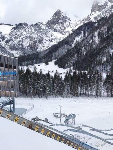 A snow-covered landscape with high mountains and a ski jump tower. In the foreground, the traces of a ski slope can be seen. | © NAZ Eisenerz