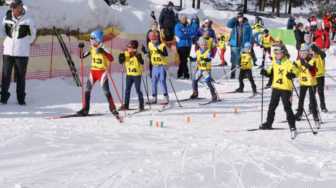 Eine Gruppe von Kindern in bunter Ski-Ausrüstung steht auf Schnee und bereitet sich auf ein Rennen vor. Zuschauer schauen auf das Event und feuern die jungen Skifahrer an. | © NAZ Eisenerz