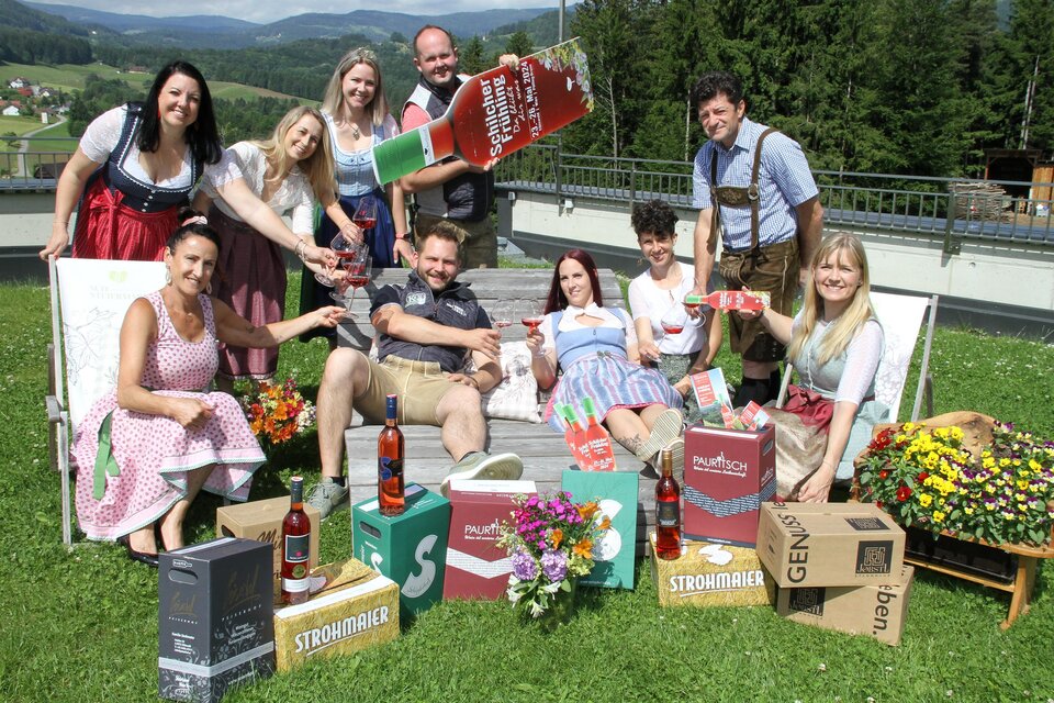 Eine fröhliche Gruppe von Menschen in traditionellen Trachten, die auf einer Wiese zusammen posieren. Sie halten Getränke und umgeben sich mit Kartons und Blumen. | © Josef Fürbass