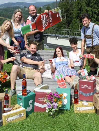 A cheerful group of people in traditional costumes posing together on a meadow. They are holding drinks and surrounded by boxes and flowers. | © Josef Fürbass