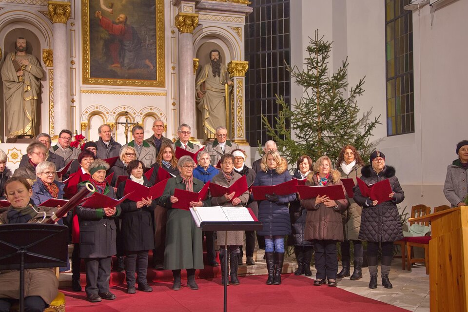 Ein gemischter Chor singt in einer Kirche, umgeben von festlicher Dekoration. Im Hintergrund steht ein Weihnachtsbaum und religiöse Statuen sind sichtbar. | © Singkreis der Volkshochschule Schladming