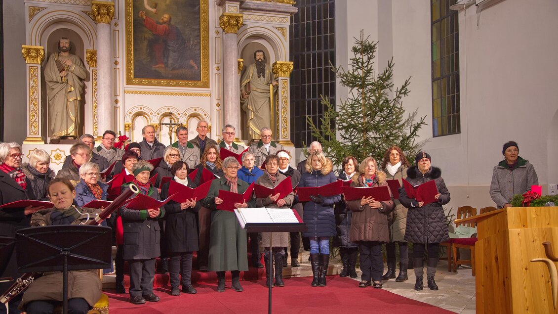 Ein gemischter Chor singt in einer Kirche, umgeben von festlicher Dekoration. Im Hintergrund steht ein Weihnachtsbaum und religiöse Statuen sind sichtbar. | © Singkreis der Volkshochschule Schladming