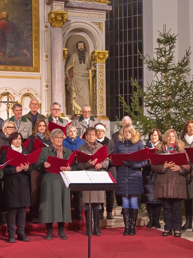 A mixed choir is singing in a church, surrounded by festive decorations. In the background, there is a Christmas tree and religious statues are visible. | © Singkreis der Volkshochschule Schladming