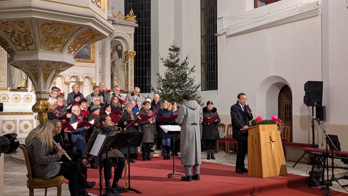 Ein Chor tritt in einer Kirche auf, umgeben von festlicher Dekoration. Vorne steht ein Redner und ein Weihnachtbaum ist zu sehen. | © Singkreis der Volkshochschule Schladming