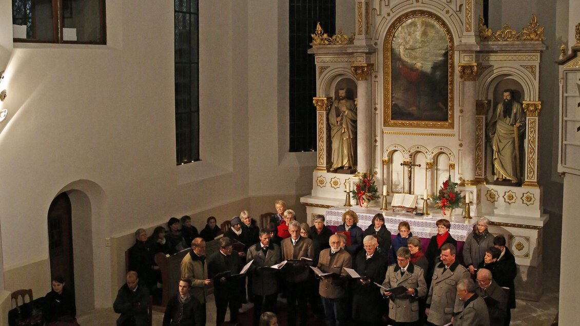 Ein Chor singt in einer Kirche mit einem schönen Altar im Hintergrund. Die Atmosphäre ist festlich und die Menschen sind in Kleidung geboten. | © photo-austria