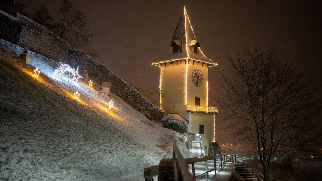 Eine verschneite Landschaft mit einem beleuchteten Turm. Im Hintergrund sind Weihnachtslichter und der Nachthimmel zu sehen.