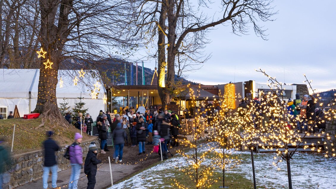 Ein festlicher Markt mit vielen Menschen und dekorativen Lichtern. Bäume und Schnee schaffen eine winterliche Atmosphäre.