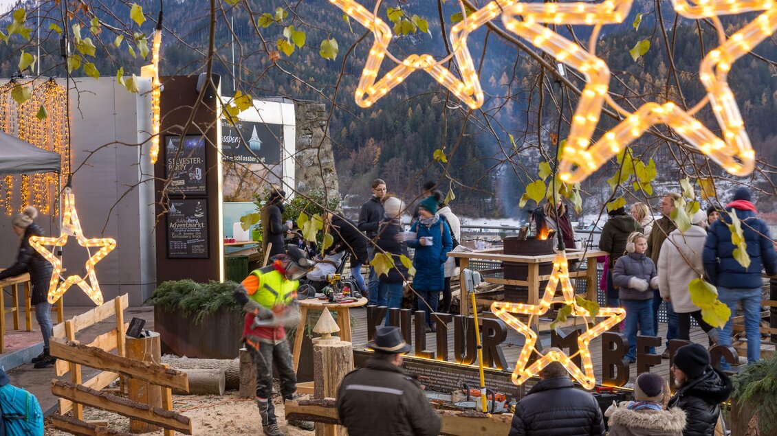 Eine festliche Szene mit leuchtenden Sternen und Menschen, die sich versammeln. Im Hintergrund sind Berge und Stände zu sehen, die eine gemütliche Atmosphäre schaffen.