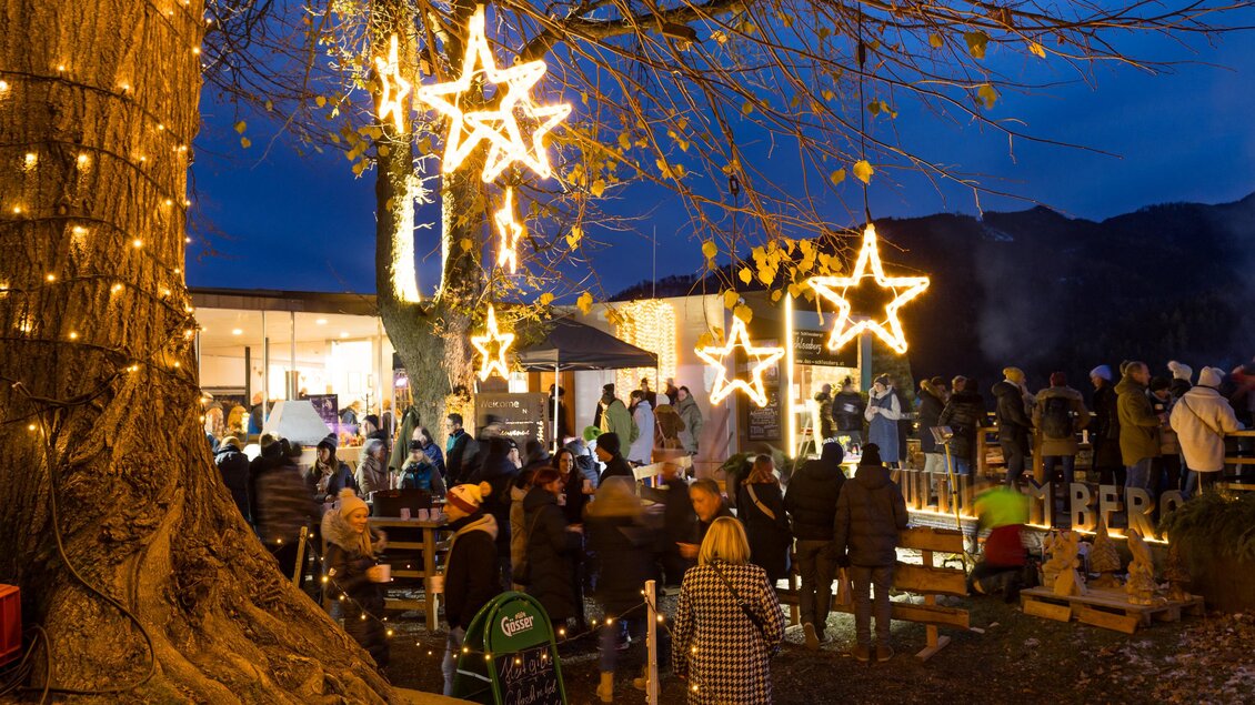 Eine festliche Szene mit leuchtenden Sternen und Menschen, die zusammen feiern. Im Hintergrund sind festlich dekorierte Stände und eine gemütliche Atmosphäre zu sehen. | © StadtBruck, Meieregger