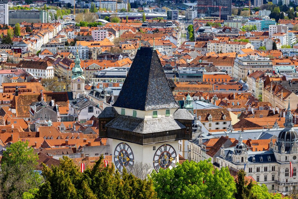 Eine Stadtansicht mit vielen roten Dächern und einem markanten Uhrturm im Vordergrund. Die Landschaft wird von grünen Bäumen umgeben und zeigt eine lebendige Stadt. | © Graz Tourismus - Harry Schiffe