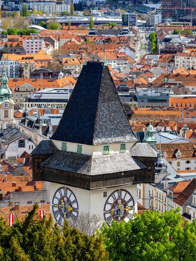 Eine Stadtansicht mit vielen roten Dächern und einem markanten Uhrturm im Vordergrund. Die Landschaft wird von grünen Bäumen umgeben und zeigt eine lebendige Stadt. | © Graz Tourismus - Harry Schiffe