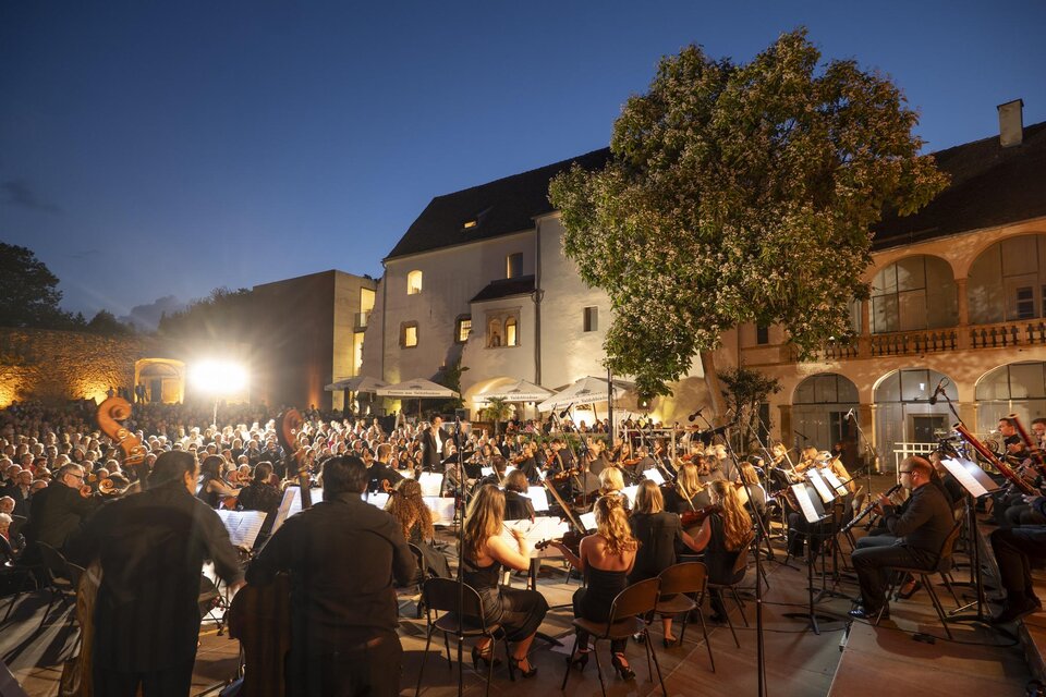 Ein großes Orchester spielt im Freien vor einem historischen Gebäude. Die Zuschauer sitzen in einem aufregenden Ambiente bei Dämmerlicht. | © Bernhard Bergmann