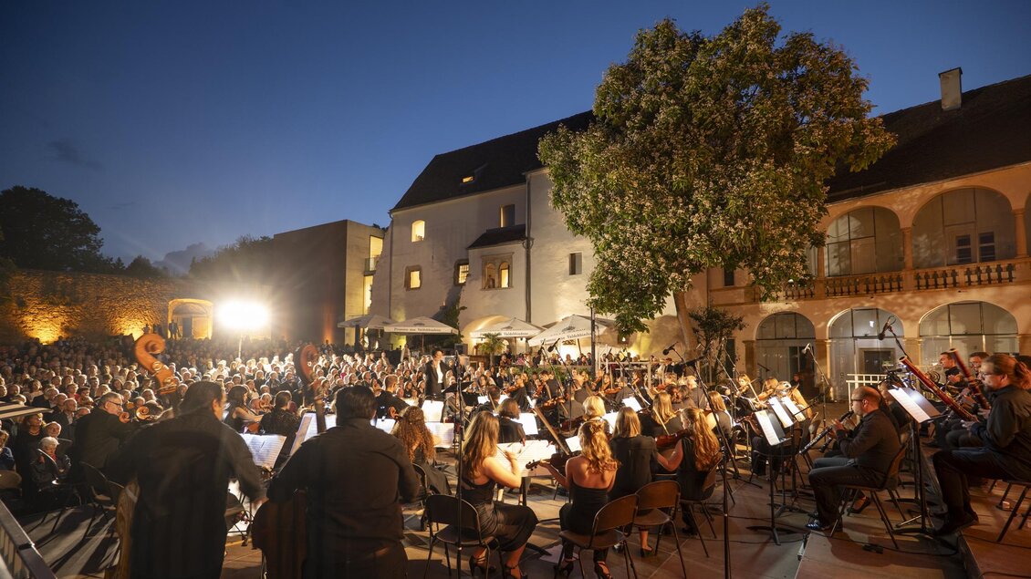 Ein großes Orchester spielt im Freien vor einem historischen Gebäude. Die Zuschauer sitzen in einem aufregenden Ambiente bei Dämmerlicht. | © Bernhard Bergmann