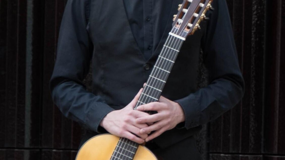 Ein Mann steht lächelnd mit einer Gitarre in der Hand. Er trägt ein schwarzes Hemd und eine Weste. | © Hermine Pack