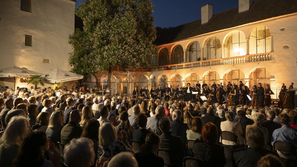 Ein Konzert im Freien mit einer großen Menschenmenge. Im Hintergrund sind historische Gebäude und Bäume unter dem Abendhimmel zu sehen.