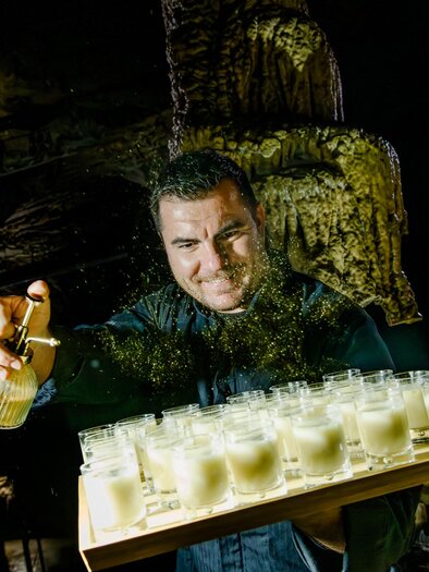A man serves drinks in a dark, impressive setting. The glasses are arranged on a tray, and the man adds a special ingredient. | © Region Graz - Mias Photoart