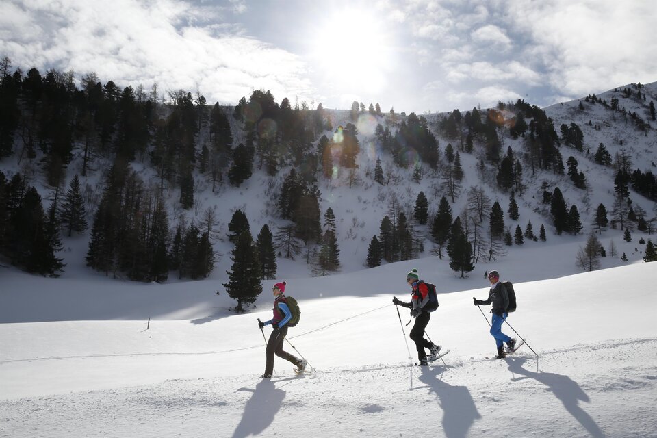 Drei Personen wandern durch eine verschneite Landschaft. Im Hintergrund sind Bäume und ein heller Himmel zu sehen. | © Herbert Raffalt