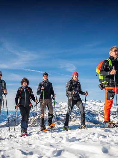 A group of five people is hiking in a snowy mountain landscape. The sky is clear and blue, and the mountains are visible in the background. | © Christoph Huber