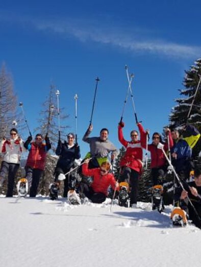 A group of people is standing on a snow-covered slope, holding ski poles in the air. The sky is blue with some clouds, and there are coniferous trees in the background. | © OCT Outdoor Consulting Group