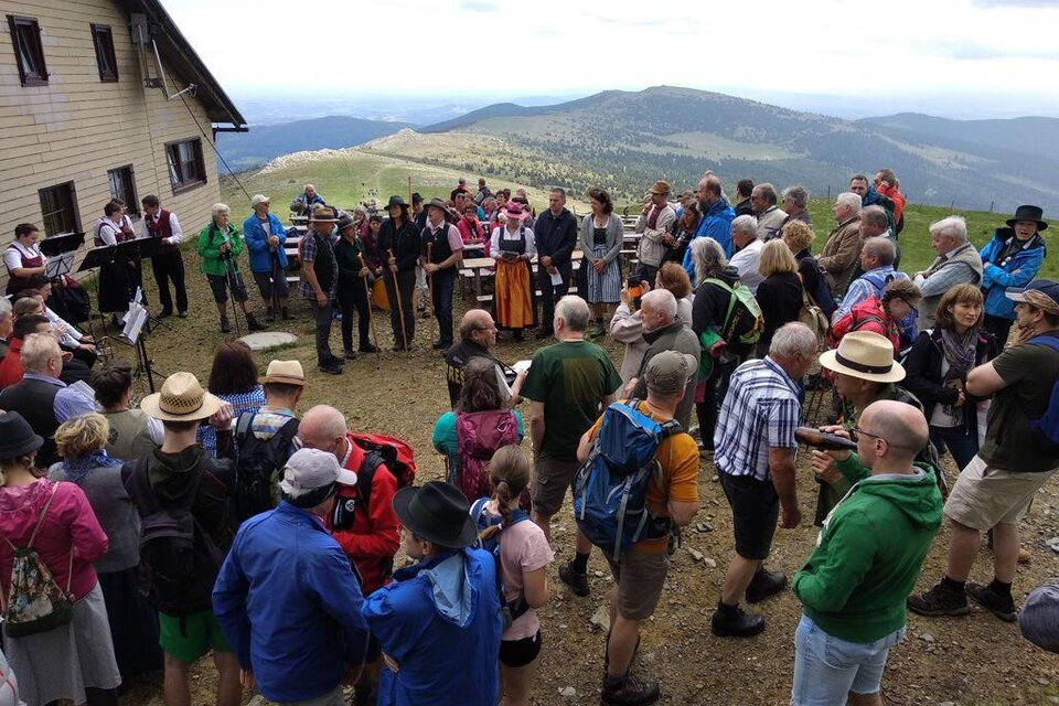 Eine Menschenmenge versammelt sich auf einem Berg, umgeben von einer schönen Landschaft. Musik wird gespielt, während die Besucher in festlicher Kleidung teilnehmen. | © Schwaigen-Reigen