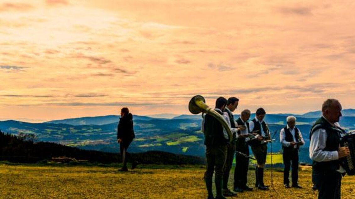 Eine Gruppe von Musikern spielt auf einem Hügel mit Blick auf eine malerische Landschaft. Der Himmel ist in warmen Farben gefärbt, während die Sonne untergeht. | © Glatzl Trahütten Alm