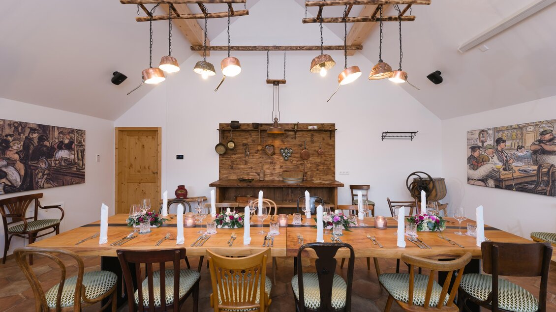 Ein rustikaler Essraum mit einem langen Holztisch und bunten Stühlen. An den Wänden hängen Bilder, und die Beleuchtung wird von hängenden Lampen bereitgestellt. | © www.zloam.at