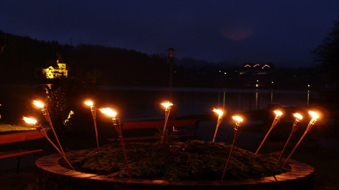 Eine romantische Nachtansicht mit Fackeln, die sanft leuchten. Im Hintergrund ist ein ruhiger See und einige Lichtpunkte von Gebäuden sichtbar. | © Gemeinde Grundlsee