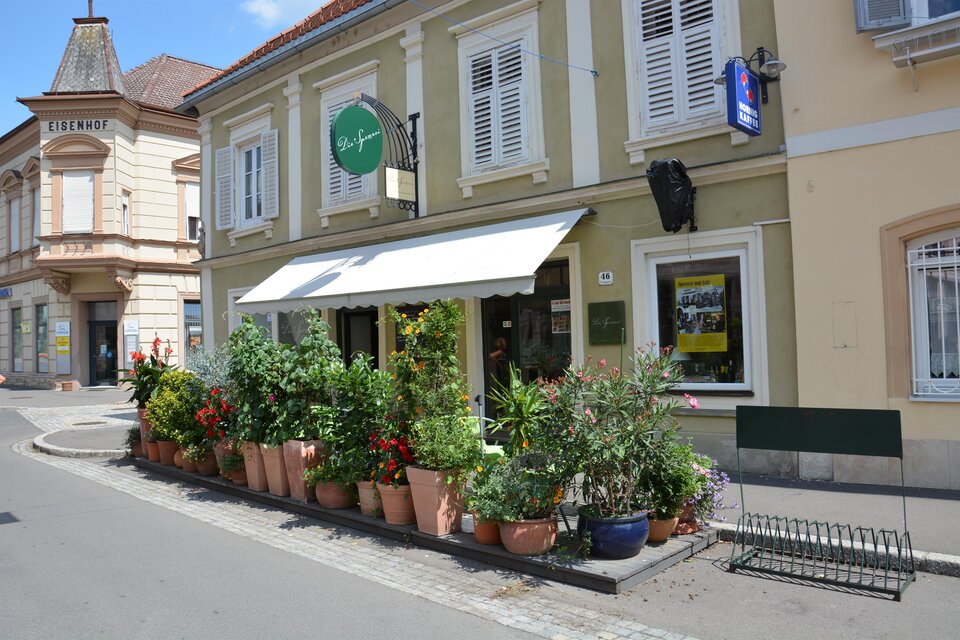Ein charmantes Restaurant mit Pflanzen in Töpfen vor der Fassade. Die Straße ist ruhig und sonnig, mit mehreren Gebäuden im Hintergrund. | © Hermann