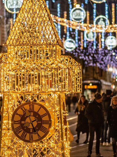 A festive street with illuminated Christmas decorations. In the foreground stands a large, sparkling clock, while people stroll by. | © achtzigzehn - Hinterleitner