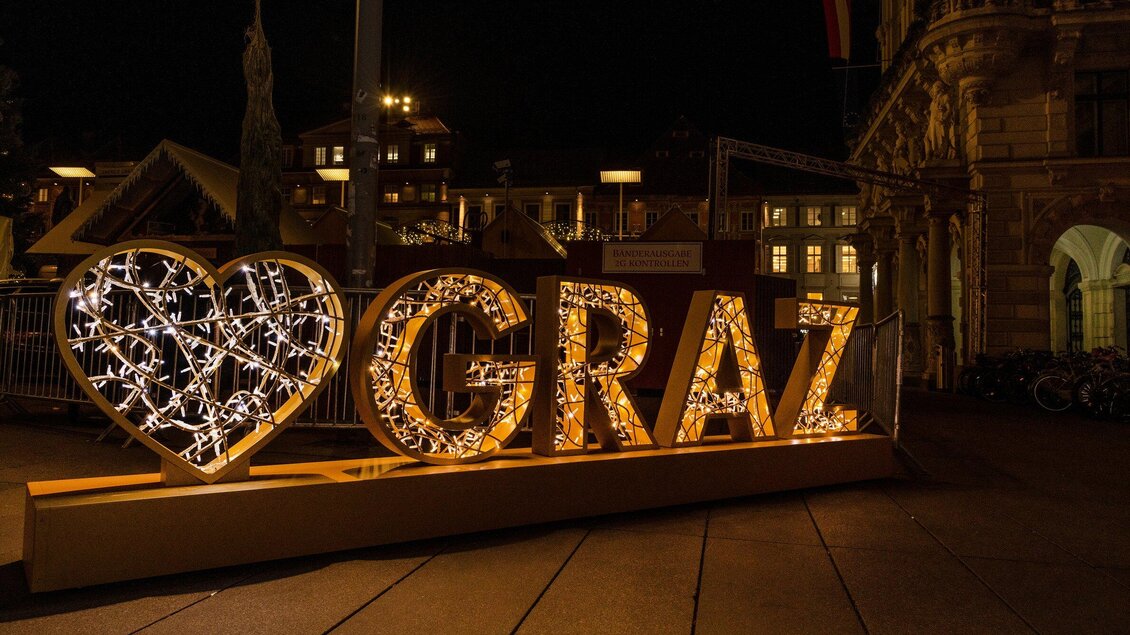 Ein beleuchtetes Herz-Symbol mit dem Schriftzug "GRAZ" steht in einer Stadtlandschaft bei Nacht. Die dekorativen Lichter schaffen eine einladende Atmosphäre. | © achtzigzehn - Hinterleitner