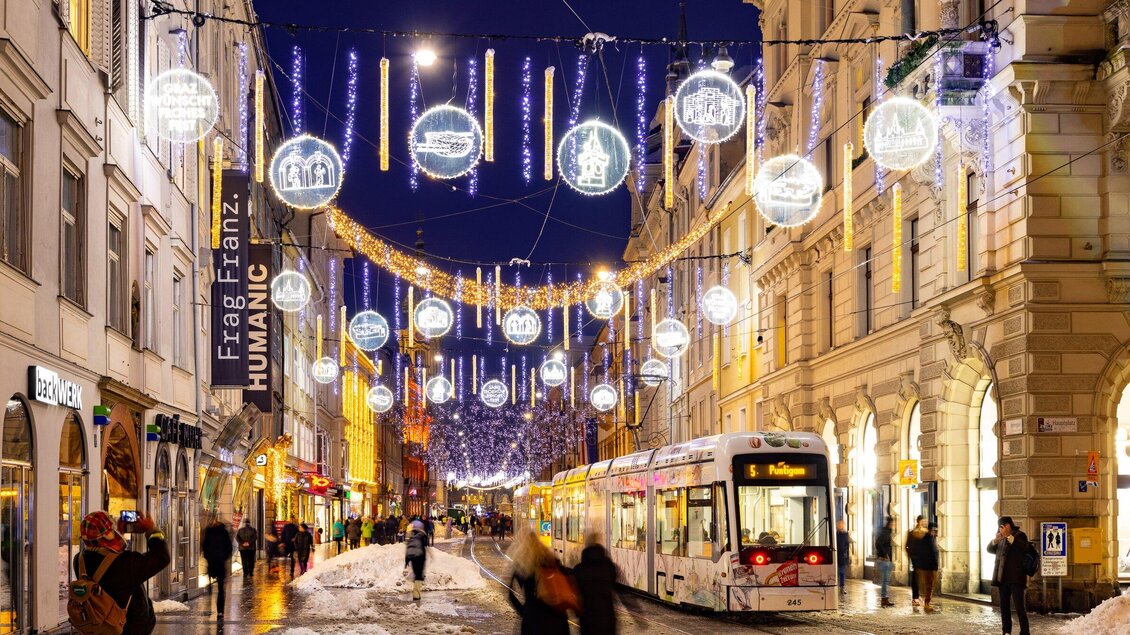 Eine festlich beleuchtete Straße mit Weihnachtsdekorationen und einem Straßenbahnwagen. Menschen flanieren durch die winterliche Szenerie. | © Graz Tourismus - Harry Schiffer