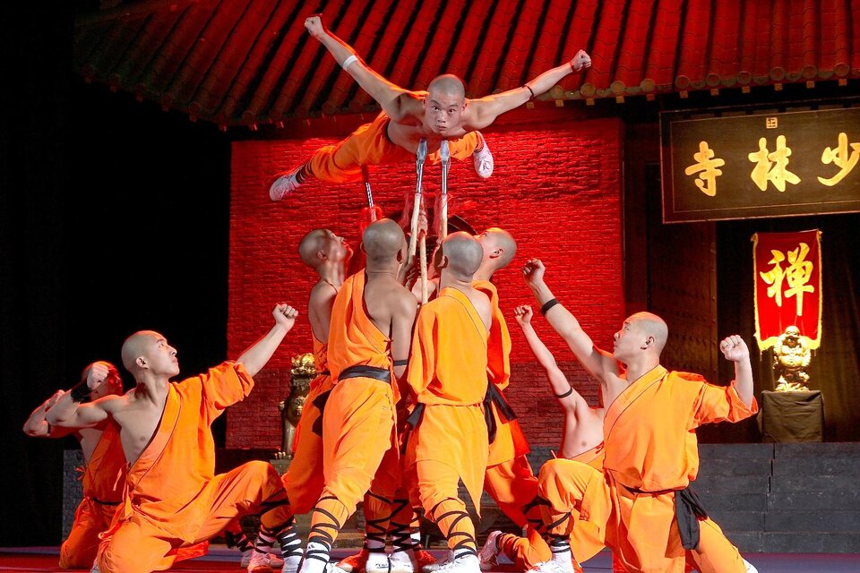 Eine Gruppe von Shaolin-Mönchen in orangefarbenen Outfits führt akrobatische Übungen auf einer Bühne vor. Im Hintergrund ist eine traditionelle chinesische Kulisse zu sehen. | ©  Pura Vida Theater und Musical GmbH