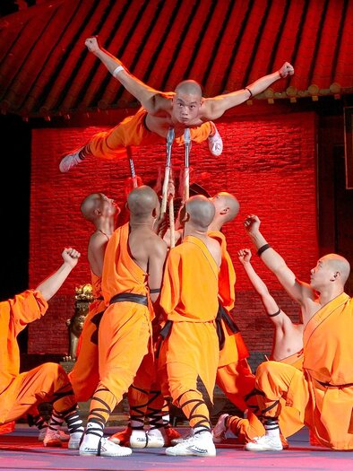A group of Shaolin monks in orange outfits performs acrobatic exercises on a stage. In the background, a traditional Chinese backdrop is visible. | ©  Pura Vida Theater und Musical GmbH