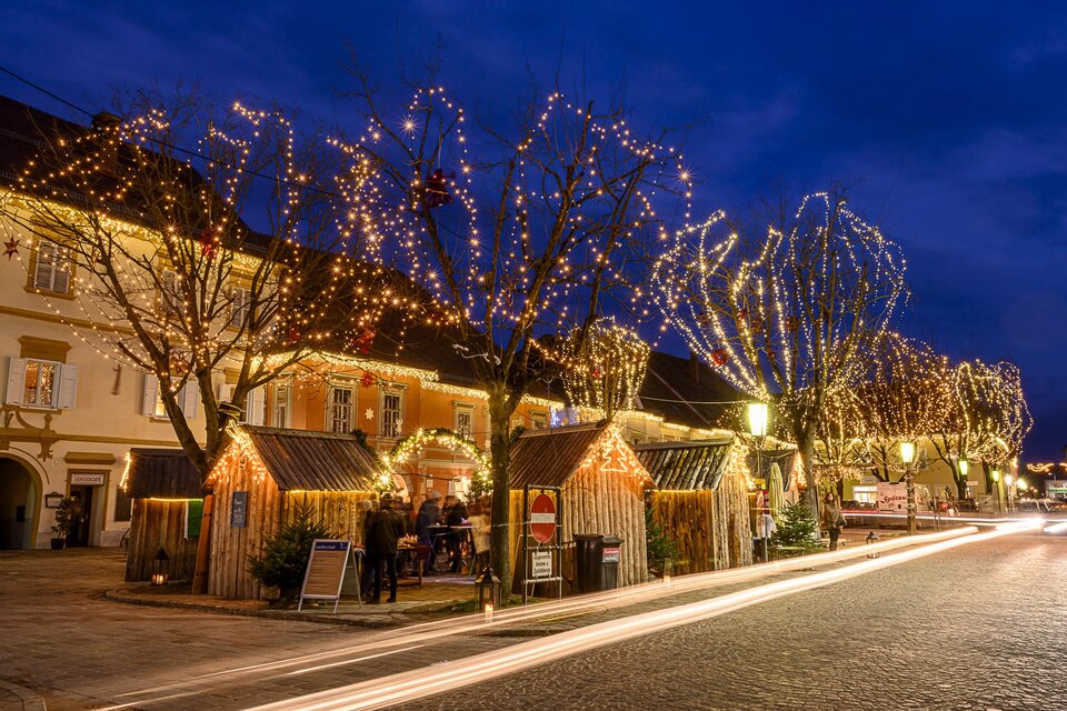 Eine festlich geschmückte Straße mit leuchtenden Bäumen und kleinen Holzhütten. Der Abendhimmel ist dunkelblau, was die Lichter noch mehr zur Geltung bringt. | © TV Thermen- & Vulkanland